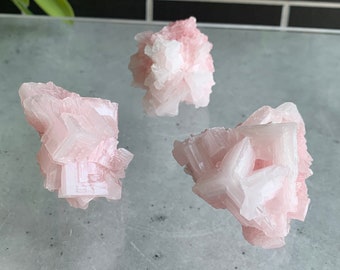 Puede incluir: Tres cristales de sal rosa del Himalaya, cada uno con una forma y textura únicas. Los cristales están agrupados en una superficie blanca.