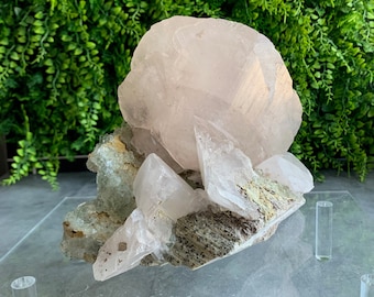 May include: A large pink crystal cluster with smaller crystals on a clear acrylic display stand. The crystal cluster is on a gray surface.