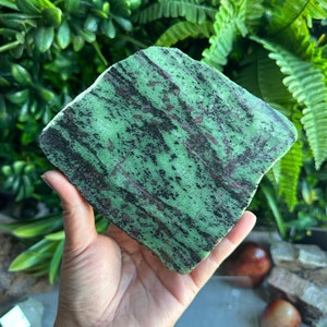 May include: A polished green and black stone with a rough, natural edge. The stone has a mottled pattern of green, black, and white.