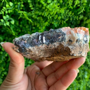 May include: A raw, natural crystal cluster with a mix of orange, white, and black minerals. The crystal has a rough, textured surface and is held in a hand.