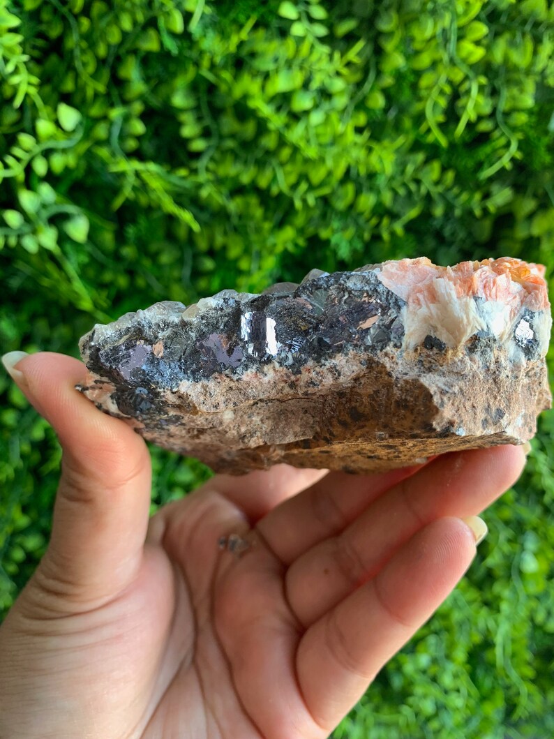 May include: A raw, natural crystal cluster with a mix of orange, white, and black minerals. The crystal has a rough, textured surface and is held in a hand.