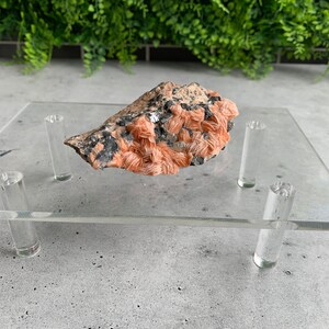 May include: A pink and white mineral specimen with a rough, natural texture. The mineral is displayed on a clear acrylic stand with four legs.
