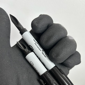 Puede incluir: Dos rotuladores permanentes Sharpie Fine Point en blanco y negro. Los rotuladores están sujetos por una mano enguantada. Los rotuladores tienen tapas negras y anillos plateados. El texto "Sharpie" y "Permanent Marker" es visible en los barriles blancos.