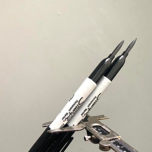 Puede incluir: Dos marcadores permanentes negros Sharpie con etiquetas blancas se están midiendo con una pinza de calibre plateado. Las etiquetas dicen "Sharpie Permanent Marker".