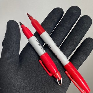 May include: Two red Sharpie fine point permanent markers with black caps. The markers are held in a black work glove.