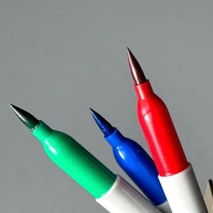 May include: Close-up of three Sharpie permanent markers in red, green, and blue, with a metal caliper. The markers have white barrels with black text and fine point tips. The red marker is in focus, with the green and blue markers slightly blurred.