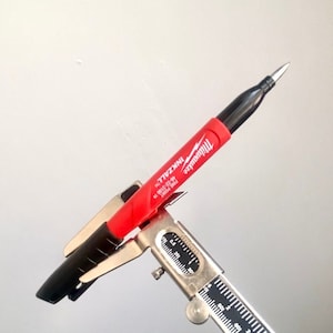 Puede incluir: Un marcador permanente de punta fina Milwaukee INKzALL rojo y negro se sostiene en una pinza de calibre plateado. El marcador tiene una tapa negra y un cuerpo rojo con el logotipo de Milwaukee y las palabras "INKzALL" y "FINE POINT" impresas en él.