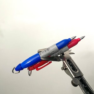 Könnte beinhalten: Ein blauer und roter Sharpie-Marker, der mit einer silbernen Schieblehre gemessen wird. Die Schieblehre zeigt eine Messung von 2,57 cm an.