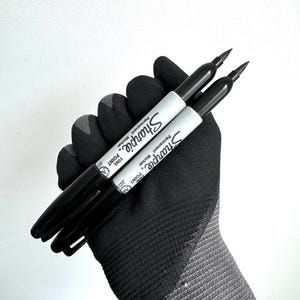 May include: Two black Sharpie permanent markers with white labels and fine point tips. The markers are held in a black and gray glove, set against a white background. The Sharpie logo is visible on the labels.