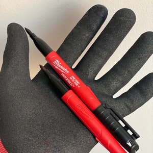 May include: Two red and black Milwaukee Inkzall fine point permanent markers. The markers are on a black work glove.