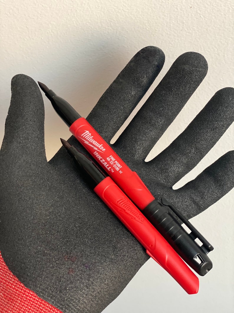 May include: Two red and black Milwaukee Inkzall fine point permanent markers. The markers are on a black work glove.