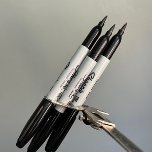 May include: Three black Sharpie fine point permanent markers are held together by a silver caliper. The markers are labeled "Sharpie Fine Point Permanent Marker" and "1-2 mm Point".