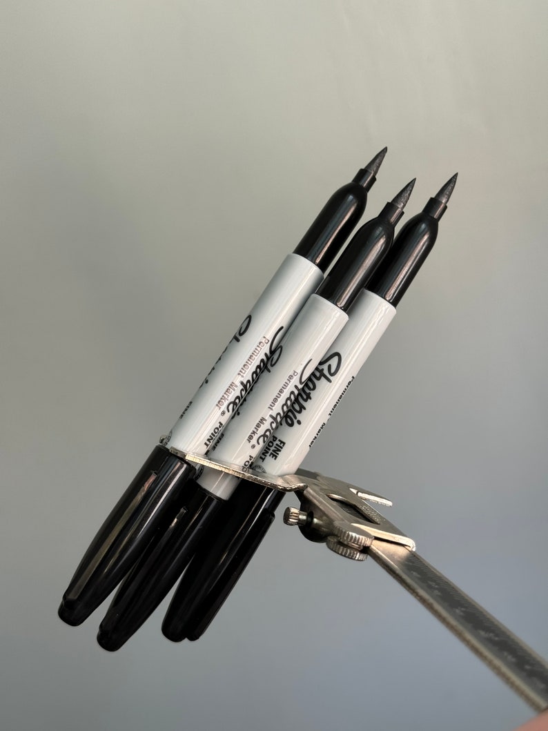 May include: Three black Sharpie fine point permanent markers are held together by a silver caliper. The markers are labeled "Sharpie Fine Point Permanent Marker" and "1-2 mm Point".