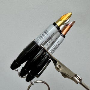 Può includere: Un set di tre pennarelli Sharpie in miniatura attaccati a un portachiavi in metallo. I pennarelli presentano fusti argentati con tappi ed estremità neri, e le punte sono in tonalità metalliche oro, argento e rame.