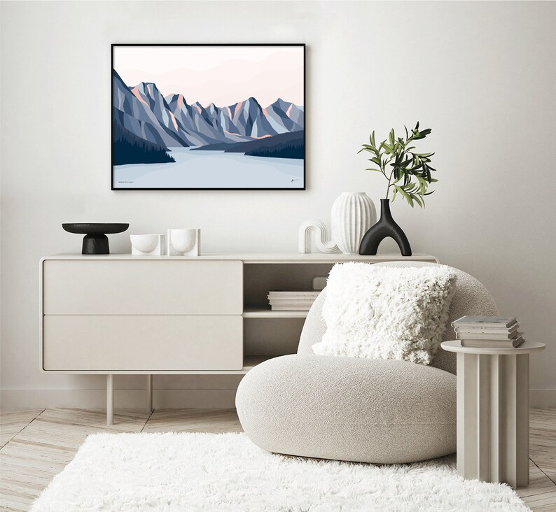 May include: A minimalist living room interior with a white armchair, a white side table, a white dresser with two drawers, and a framed print of a mountain landscape with a lake in the foreground. The print is in shades of blue and pink.