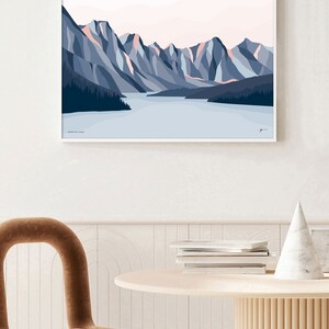 May include: A framed illustration of a mountain range with a lake in the foreground. The mountains are a mix of blue and pink hues, with a light blue sky above. The lake is a light blue color, and there are trees on the shore.
