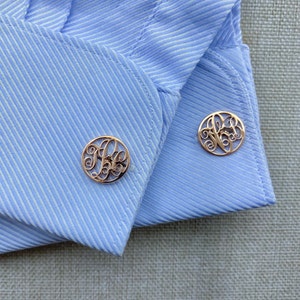 Può includere: Un paio di gemelli d'oro con un design a monogramma. I gemelli sono su una camicia a righe blu chiaro.