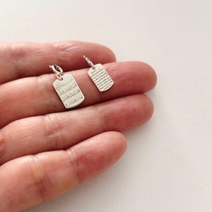 Puede incluir: Dos pequeños colgantes rectangulares plateados con esquinas redondeadas. Cada colgante tiene un pequeño lazo en la parte superior para colgar. Los colgantes están grabados con texto. Los colgantes se muestran sobre un fondo claro.