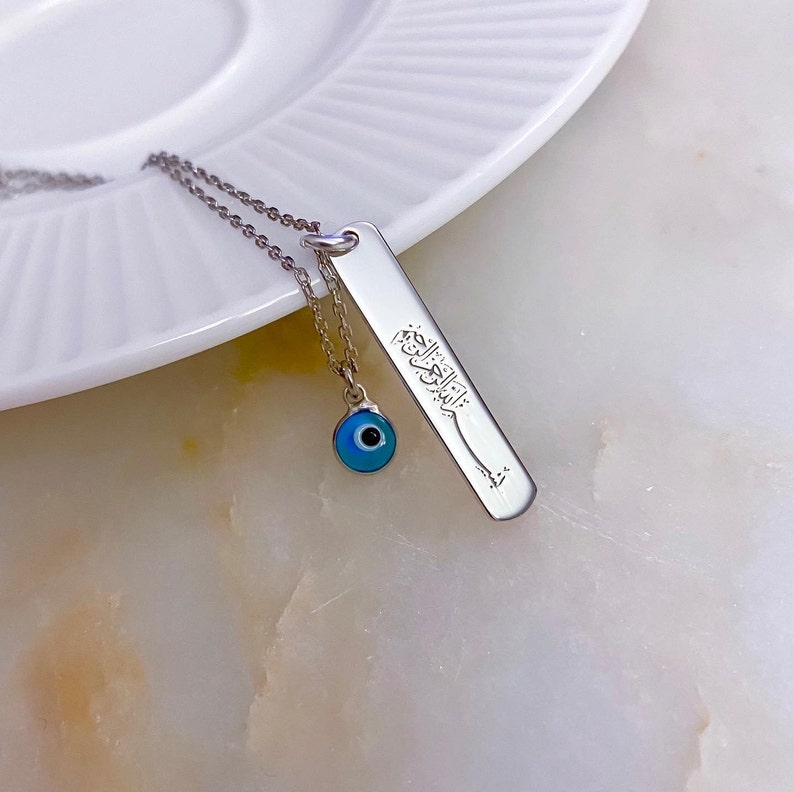 May include: Silver necklace with a blue evil eye charm and a pendant with Arabic calligraphy.