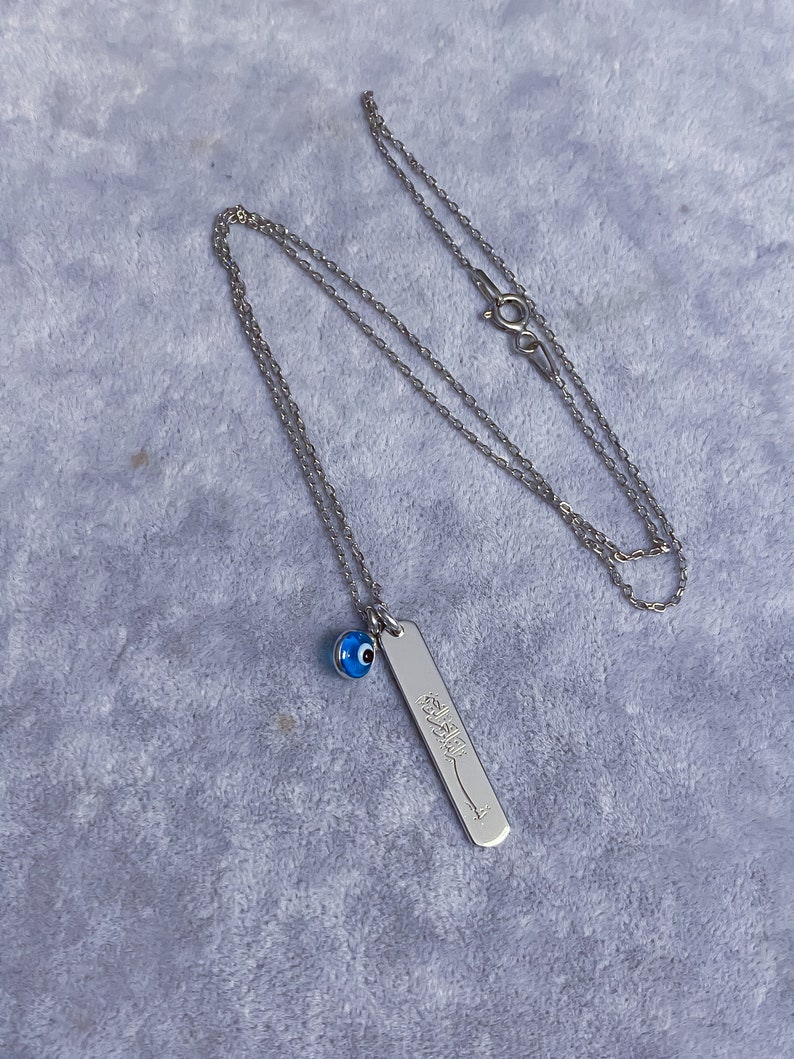 May include: Silver chain necklace with a blue evil eye charm and a silver pendant with Arabic calligraphy.