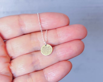 May include: A delicate silver chain necklace featuring a small, circular gold-toned pendant. The pendant is engraved with multiple lines of fine, dark text that covers the entire surface of the round metal disc, suspended from a thin, polished silver link chain.