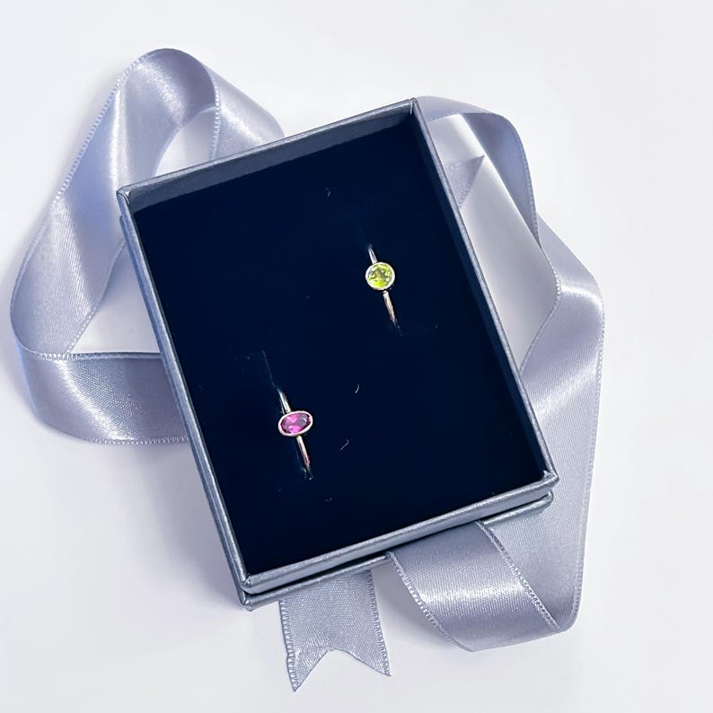 Pu&ograve; includere: Due anelli in una scatola nera con un nastro grigio. Un anello ha una pietra ovale rosa e l'altro ha una pietra ovale verde.