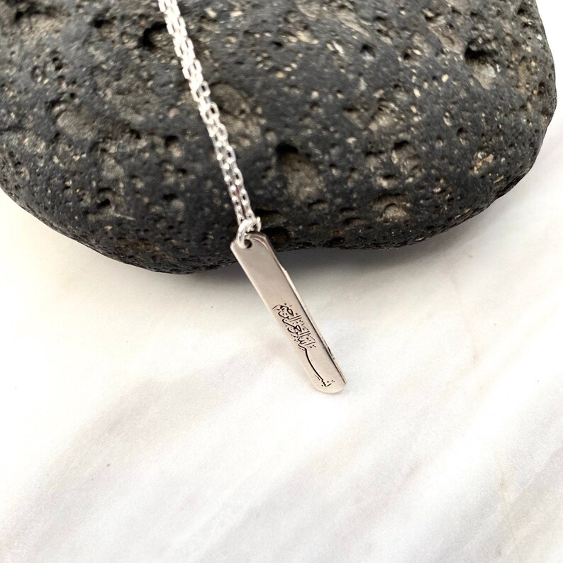May include: A silver necklace with a rectangular pendant featuring Arabic calligraphy. The pendant is hanging from a delicate silver chain.