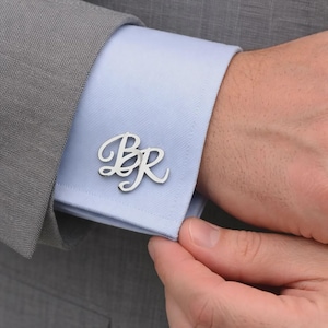 Op de afbeelding: Een zilverkleurige manchetknop met de gestileerde cursieve letters B en R, bevestigd aan de manchet van een lichtblauw overhemd onder een grijze blazer. De letters zijn verbonden in een vloeiend ontwerp met een gepolijste afwerking.