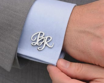 Puede incluir: Un gemelo de metal plateado con las letras cursivas estilizadas B y R, sujeto al puño de una camisa de vestir azul claro bajo una americana gris. Las letras están conectadas en un diseño de escritura fluida con un acabado pulido.