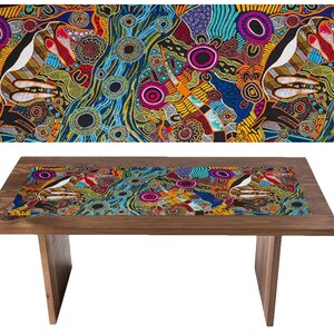 Op de afbeelding: Een houten tafel met een kleurrijke tafelrunner met een Aboriginal kunstontwerp. De runner heeft een blauw, geel, rood en zwart patroon met cirkels en lijnen.