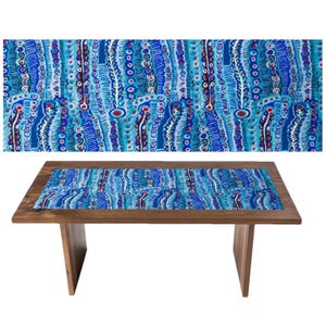 May include: A wooden table with a blue patterned table runner. The runner features a repeating design of vertical lines in shades of blue, white, and red. The table has a rectangular top and angled legs, and is set against a white background.
