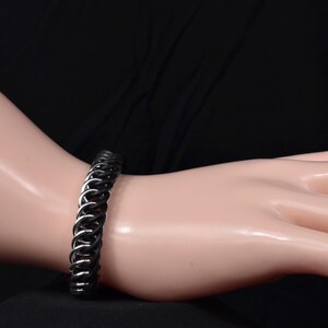 May include: A black and silver chainmail bracelet. The bracelet is made of interlocking rings, with alternating black and silver links. The bracelet is shown on a light-skinned arm against a black background. The bracelet is a piece of jewellery.