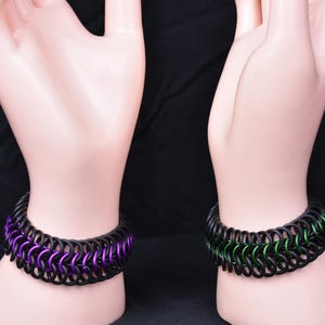 May include: Two black metal bracelets with a chainmail design. One bracelet has purple accents and the other has green accents.