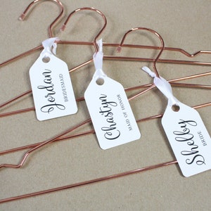 May include: Three rose gold hangers with white tags attached. The tags are personalized with the names "Jordan", "Chastyn", and "Shelby" and their corresponding titles: "Bridesmaid", "Maid of Honor", and "Bride".