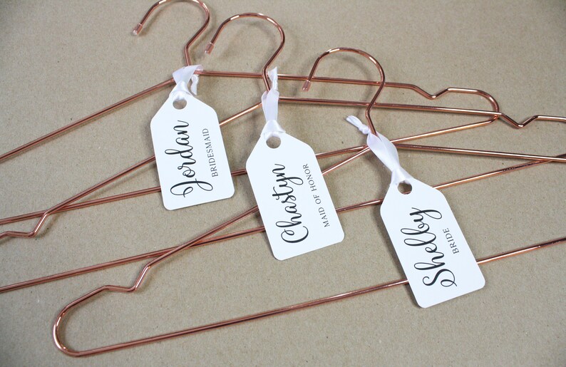 May include: Three rose gold hangers with white tags attached. The tags are personalized with the names "Jordan", "Chastyn", and "Shelby" and their corresponding titles: "Bridesmaid", "Maid of Honor", and "Bride".