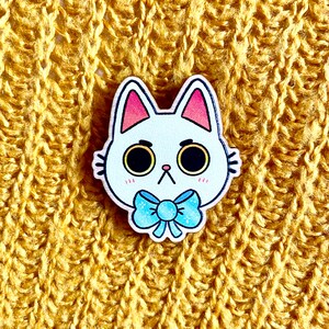 May include: White wooden cat pin with blue bow tie. The cat has large eyes and a cute expression.