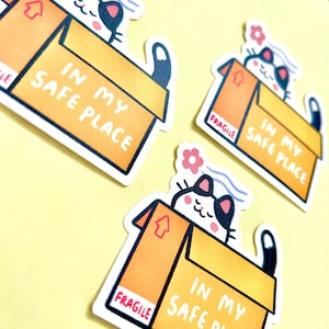 May include: Three die-cut stickers featuring a black and white cat sitting inside a brown cardboard box. The box has the text IN MY SAFE PLACE and a red FRAGILE label. A small pink flower and blue wavy lines appear above the cat's head.