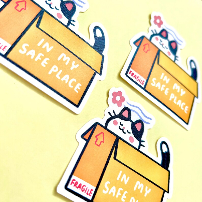 May include: Three die-cut stickers featuring a black and white cat sitting inside a brown cardboard box. The box has the text IN MY SAFE PLACE and a red FRAGILE label. A small pink flower and blue wavy lines appear above the cat's head.