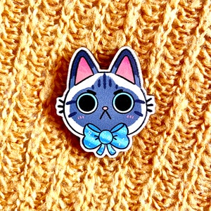 May include: A white and gray cat with blue eyes wearing a blue bow tie. The cat is cartoon-style and has a surprised expression. The cat is on a yellow knitted background.