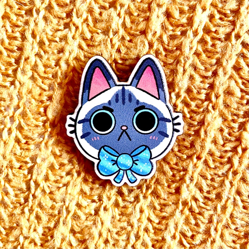May include: A white and gray cat with blue eyes wearing a blue bow tie. The cat is cartoon-style and has a surprised expression. The cat is on a yellow knitted background.