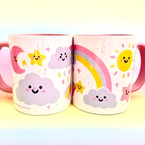 May include: Two white ceramic mugs with pink handles and rims. Each mug features a whimsical design of a smiling sun, moon, stars, clouds, and a rainbow in pastel colours. The mugs are decorated with a cute illustration.