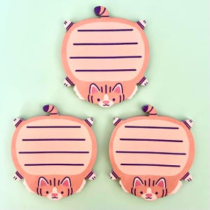 May include: Three cat-shaped sticky notes. Each note is a rounded rectangle with a pink background and a cat face at the bottom. The notes have purple stripes for writing and a purple tail. The background is a light green.