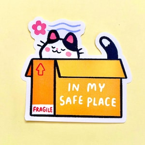 May include: A die-cut sticker featuring a black and white cat with pink cheeks peeking out of an orange cardboard box. The box has an upward-pointing arrow, the word FRAGILE, and the text IN MY SAFE PLACE. A small pink flower and blue wind lines are above the cat.