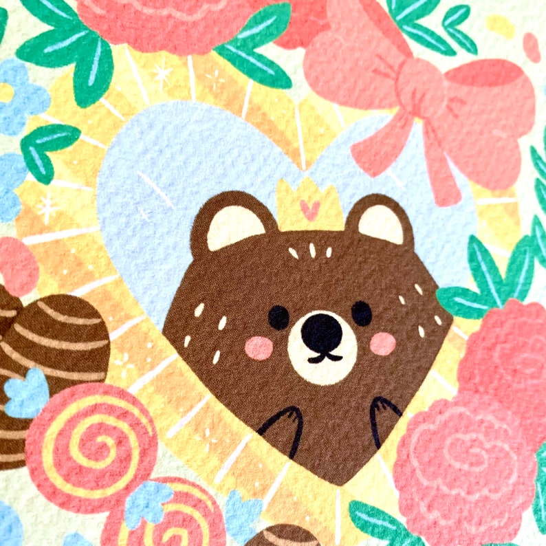 May include: A brown bear with a crown and pink cheeks is inside a heart-shaped frame. The frame is surrounded by flowers and a pink bow.