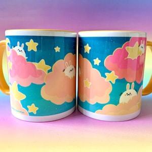 May include: Two yellow-rimmed ceramic mugs with a blue night sky background. The mugs feature a repeating pattern of white bunnies sleeping on pink and yellow clouds with yellow stars. The text "Sleepy Illustration" is printed on the bottom of one mug.