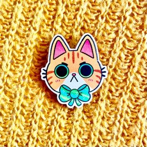 May include: A wooden pin of a cartoon cat with orange and white fur, blue eyes, and a turquoise bow tie. The cat has a grumpy expression.