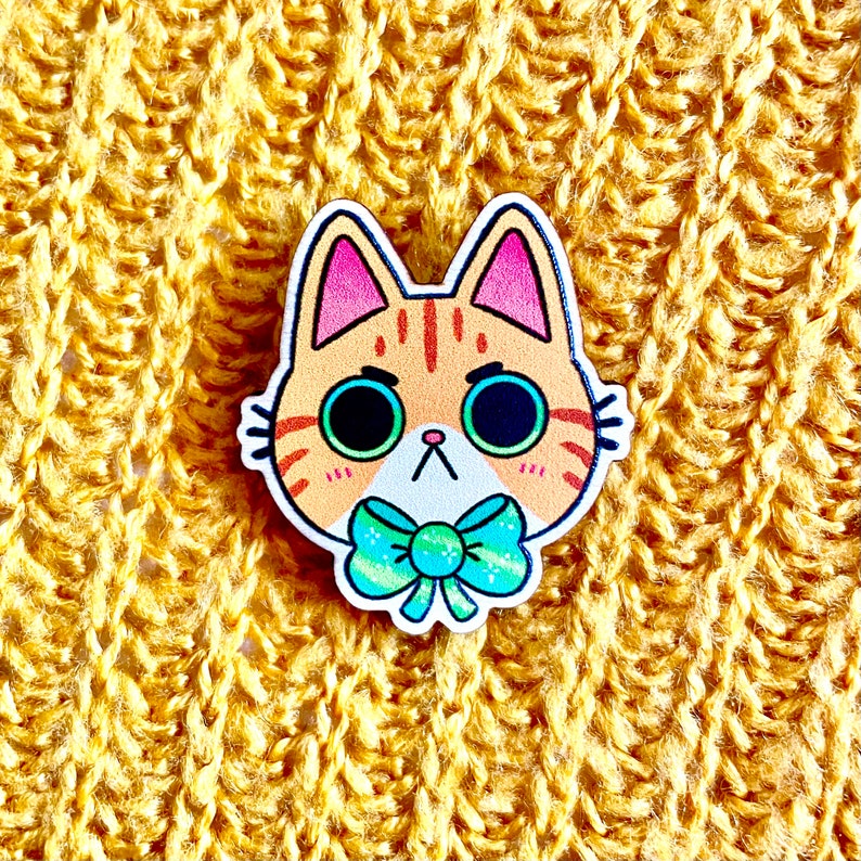 May include: A wooden pin of a cartoon cat with orange and white fur, blue eyes, and a turquoise bow tie. The cat has a grumpy expression.