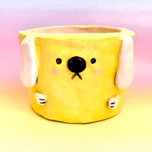 May include: A yellow ceramic planter shaped like a dog with white ears and black eyes. The planter has a small black nose and a mouth with a slight frown.