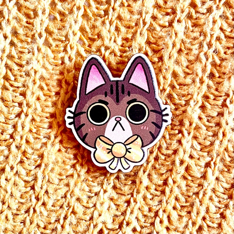 May include: A brown and black cat with a yellow bow tie. The cat has a grumpy expression on its face. The cat is made of wood and is a pin.