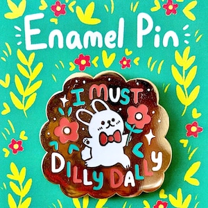 Può includere: Spilla smaltata con bordo dorato e un coniglietto bianco che tiene un fiore. La spilla ha il testo "I MUST DILLY DALLY" in rosso e verde acqua. Lo sfondo è verde acqua con accenti floreali gialli e rossi.